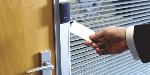 A hand presenting a white access card to a wall mounted reader beside a timber door fitted with a lever handle